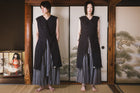 Person wearing a black sleeveless top and gray skirt standing in front of a wooden screen with traditional Japanese artwork.