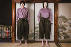 Person wearing a purple top and brown pants standing in front of a wooden screen with a painting.