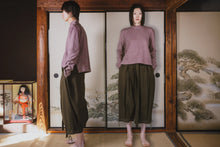 Person wearing a purple shirt and green pants standing in front of a traditional Japanese screen with a doll display.