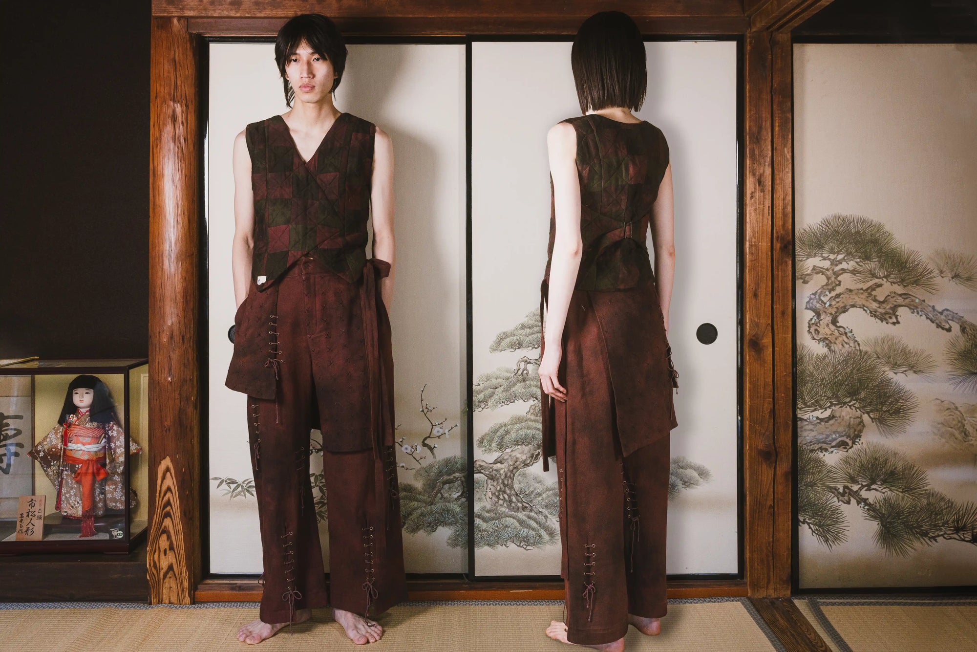 Person wearing a brown outfit standing in front of a traditional Japanese screen with a doll and artwork on the side.