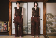Two people wearing matching brown outfits standing in front of a traditional Japanese screen with nature scenes.
